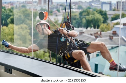 Industrial Mountaineering Cleaning Service Worker Hanging On Rope And Wiping Window With Microfiber Cloth. Cleaner Using Safety Lifting Equipment While Cleaning Glass Of High-rise Building.