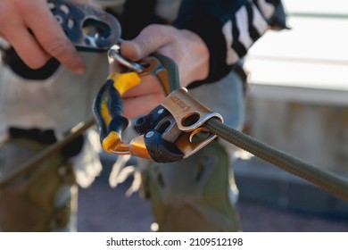 Industrial Mountaineering Equipment, Rope And Descender For Ascent In A Curved State. Background Image Of The Process Of Work On A High-rise Building. Unset Light On Metal Climbing Gear