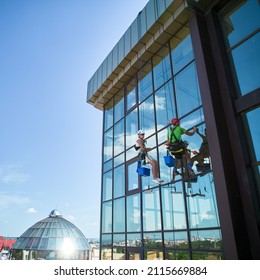 Industrial Mountaineering. Male Washers Hanging On Ropes And Using Cleaning Tools While Washing Building Windows. Two Men Window Cleaners In Safety Helmets Working Together At High-rise Building.