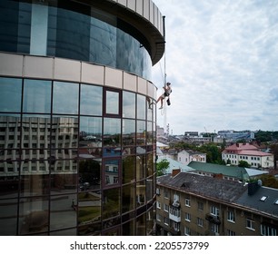 Industrial Mountaineering Professional Cleaner Hanging On Climbing Rope And Cleaning Window Of High-rise Building. Man Window Washer Using Safety Lifting Equipment While Washing Skyscraper Facade.