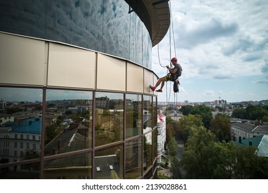 Industrial Mountaineering Worker Cleaning Glass Window Of High-rise Building. Male Cleaner Using Safety Lifting Equipment While Washing Window Of Skyscraper.