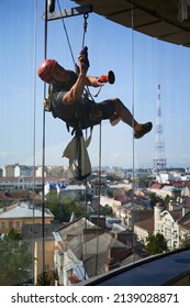 Industrial Mountaineering Worker Hanging On Climbing Rope And Cleaning Window Of High-rise Building. Man Using Safety Lifting Equipment While Wiping Skyscraper Window With Rag.