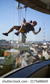 Industrial Mountaineering Worker Hanging On Climbing Rope And Cleaning Window Of High-rise Building. Man Using Safety Lifting Equipment While Wiping Skyscraper Window With Rag.