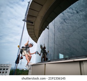 Industrial Mountaineering Worker Professional Cleaner Hanging On Climbing Rope And Cleaning Window Of High-rise Building. Window Washer Using Safety Lifting Equipment While Washing Skyscraper Facade