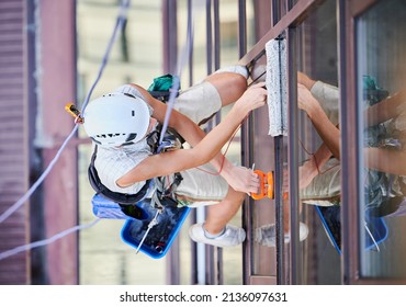 Industrial Mountaineering Worker Washing Glass Windows Of High-rise Building, Hanging On Safety Climbing Rope. Man Window Cleaner In Protective Helmet Cleaning Skyscraper Facade. Top View.