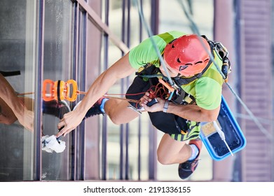 Industrial Mountaineering Worker Washing Glass Windows Of High-rise Building, Hanging On Safety Climbing Rope. Man Window Cleaner In Protective Helmet Cleaning Skyscraper Facade. Top View.