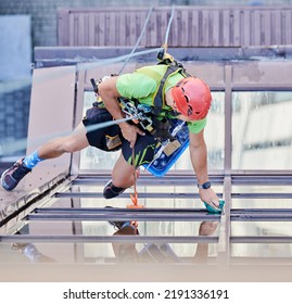 Industrial Mountaineering Worker Washing Glass Windows Of High-rise Building, Hanging On Safety Climbing Rope. Man Window Cleaner In Protective Helmet Cleaning Skyscraper Facade. Top View.