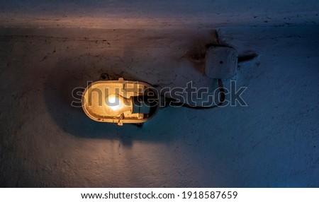 Industrial old ceiling lamp (storage or factory lamp) with vintage switched on bulb on dirty wall in a dark warehouse, basement or attic
