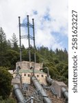 Industrial penstocks and tall exhaust towers at the Pit 3 Powerhouse, a hydroelectric facility in Shasta County, California, surrounded by forested hills.