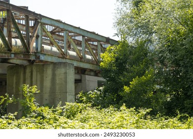 An industrial pipe crossing a stream with a rusty bridge and trees. - Powered by Shutterstock