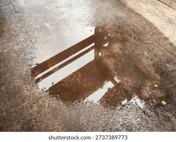 Industrial pipe structure reflected in a puddle of water on wet asphalt surface. The image captures an abstract reflection of metal framework distorted by ripples and uneven pavement texture. Moist ground and reflective water create a gritty urban atmosphere symbolizing infrastructure, industrial environment, and post-rain conditions. The composition blends texture, reflection, and geometry, making it suitable for conceptual backgrounds, urban themes, and industrial design visuals.