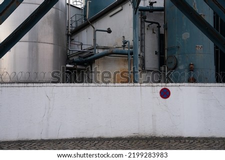 Industrial piping of a chemical plant. Painted in turquoise. Rusty steel construction, barbed wire and a white painted wall with traffic sign 