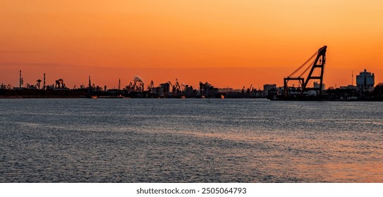 Industrial port silhouette along a tranquil water's edge, under a vivid orange sunset sky, punctuated by cranes and smoke rising in the distance. - Powered by Shutterstock