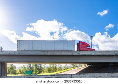 Industrial powerful red big rig classic semi truck transporting commercial cargo in dry van semi trailer running on the concrete bridge across the wide highway road on the cloud sky background