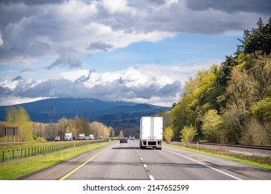 Industrial professional long hauler white big rig semi truck transporting commercial cargo in dry van semi trailer running on the wide highway road with opposite traffic and forest on the sides