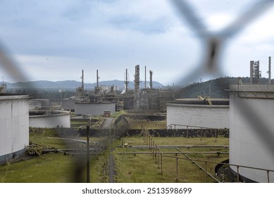 Industrial refinery complex, multiple large structures, including storage tanks and towers, view partially obscured by wire fence, highlighting security or restricted access, processing raw materials