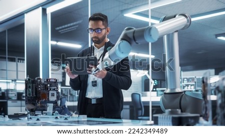Industrial Robotics Professional Interacting with Robotic Arm During a Research Phase in a High Tech Startup. Scientist Uses Tablet Computer to Manipulate and Program the Robot to Move a Microchip.