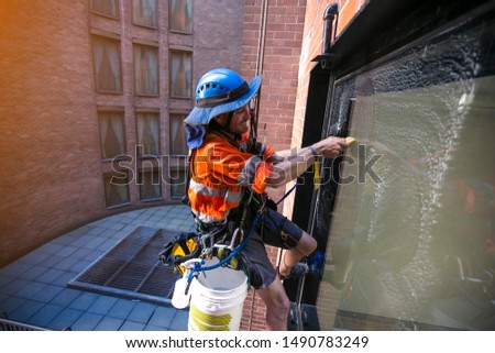 Industrial rope access abseiler window cleaner wearing full safety harness, helmet fall protection abseiling with twin low stretch ropes conducting cleaning window with yellow scraper Sydney CBD  