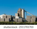 An industrial site with a tall silo and conveyor, surrounded by stacked concrete blocks, under a bright blue sky.