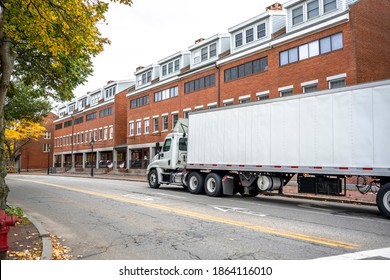 Industrial White Big Rig Day Cab Semi Truck Tractor With Roof Spoiler Transporting Commercial Cargo In Dry Van Semi Trailer Moving On The Small City Street Road With Buildings In New England