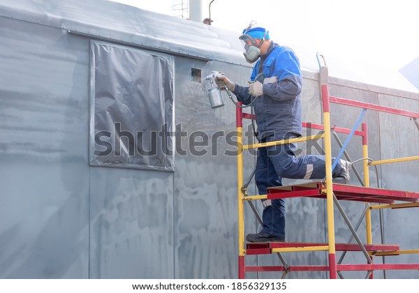 Industrial work. Priming of metal products\
from the compressor gun. A worker in overalls and a respirator\
paints the body of a truck trailer or a metal\
car.