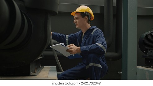 Industrial worker in blue overalls and a yellow hard hat attentively examines machinery with clipboard in hand. - Powered by Shutterstock