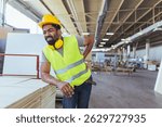 An industrial worker in a factory experiencing back pain, emphasizing occupational health and safety awareness. The worker wears a hard hat and reflective vest while protecting himself  