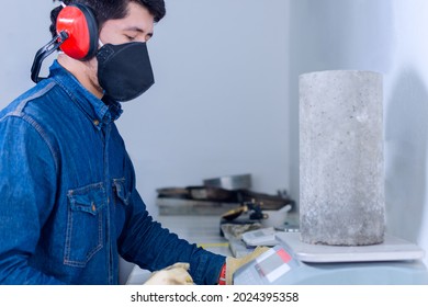 Industrial Worker Man With Protective Mask And Hearing Protectors Holding A Concrete Cylinder On A Scale