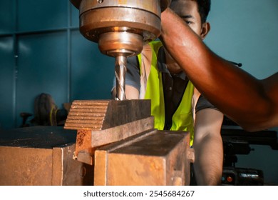 An industrial worker operating a drill press in a workshop. The focus is on the precision and concentration required to handle machinery. The worker wears a safety vest, ensuring a secure work. - Powered by Shutterstock