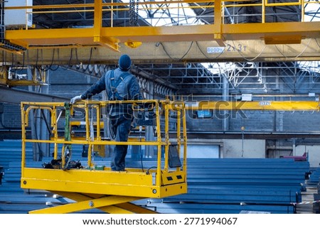 Industrial worker standing on elevated yellow scissor lift platform inside warehouse with overhead crane systems and stacked steel materials