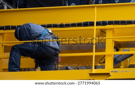 Industrial worker using an angle grinder while performing maintenance on a yellow overhead crane platform inside a large steel warehouse with visible sparks and safety harness equipment.