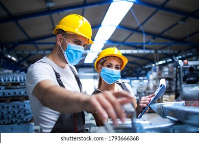 Industrial Workers With Face Masks Protected Against Corona Virus Discussing About Metal Parts In Factory. People Working During COVID-19 Pandemic.