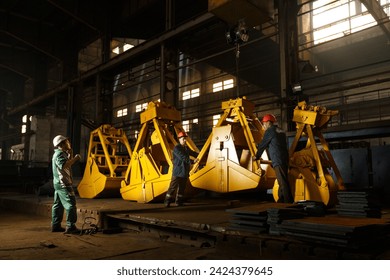 Industrial workers inspect yellow grapples in factory setting. Heavy equipment manufacturing process, teamwork with safety helmets. Quality control in metal fabrication, engineered machinery parts. - Powered by Shutterstock