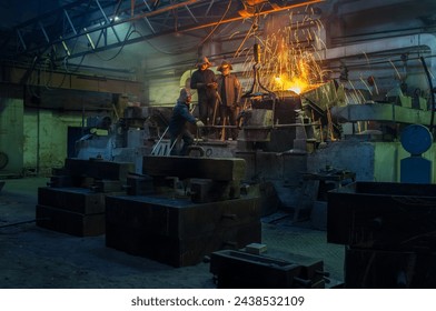 Industrial workers in protective gear casting metal in foundry. Team supervises molten steel pouring, safety helmets, heavy machinery. Manufacture, teamwork, metallurgy, high temperature workshop. - Powered by Shutterstock