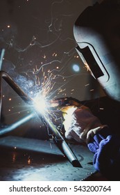 Industrial Workshop Man Welding Round Pipe On A Metal Work Table, Colorful Smoke, Sparks And Reflections