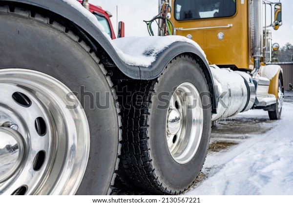 Industrial yellow professional day cab big rig semi\
truck tractor with twin axles and doubled wheels and tires with a\
large tread standing on the winter parking lot with snow and ice on\
car and lot