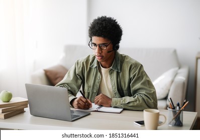 Industrious Black Teenager In Headset Studying Remotely From Home, Writing In Notebook. African American Student Taking Online Class On Laptop, Watching Video Lesson, Participating In Conference