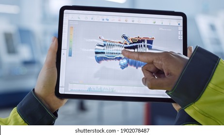 Industry 4.0 Factory: Chief Engineer And Project Supervisor In Safety Vests And Hard Hats, Talk, Use Digital Tablet Computer, Screen Shows 3D Concept Of New Jet Engine. Workshop With Machinery.