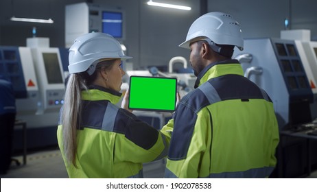 Industry 4.0 Factory: Chief Engineer And Project Supervisor In Safety Vests And Hard Hats, Talk, Use Digital Tablet Computer With Green Screen, Chroma Key. Workshop With Machinery.