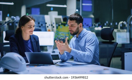 Industry 4.0 Modern Factory Meeting Room: Chief Engineer Holds Mechanism, Shows It To Female Designer, Use Laptop. Scientists In Contemporary Lab Build Electronic Machinery For With Futuristic Design