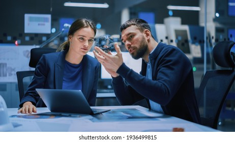 Industry 4.0 Modern Factory Meeting Room: Chief Engineer Holds Mechanism, Shows It To Female Designer, Use Laptop. Scientists In Contemporary Lab Build Electronic Machinery For With Futuristic Design