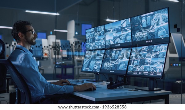 Industry 4.0\
Modern Factory: Security Operator Controls Proper Functioning of\
Workshop Production Line, Uses Computer with Screens Showing\
Surveillance Camera Feed. High-Tech\
Security