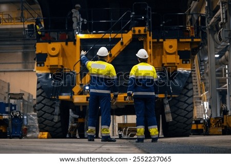 Industry big yellow mining truck maintenance. Two engineers control repair of dump car in garage of factory.