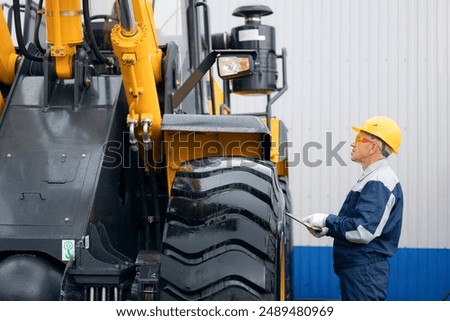 Industry engineer uses computer tablet for control monitoring condition of industrial equipment. Adult man in uniform and yellow helmet against background of mining tractor.