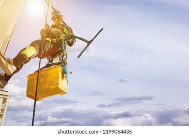 Industry Urban Works. Close Up Industrial Mountaineering Worker In Uniform Hangs Over Residential Building At Sky Background, Washing Exterior Facade. Rope Access Laborer On Wall Of House. Copy Space