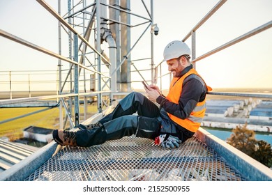 An industry worker sitting on metal construction on a break and scrolling on the phone.
