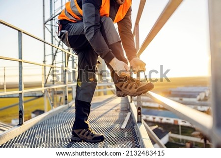 An industry worker tying shoelace on work shoes while standing on metal construction.
