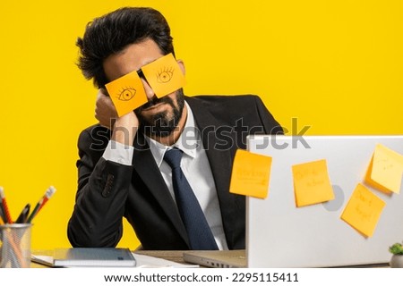 Inefficient tired young businessman working sleeping on laptop computer with eyes stickers on face at office workplace desk. Indian lazy manager freelancer man on yellow background. Cheating to sleep