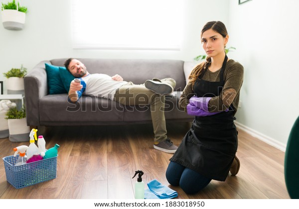 Inequality concept. Portrait of angry woman doing all\
the cleaning and chores while partner is lying on the couch and\
watches TV
