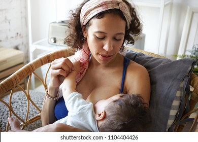 Infancy, motherhood, nutrition, lactation and breastfeeding concept. Indoor portrait of tender attractive young mom nursing her baby boy, giving him all love and affection, sitting in bedroom